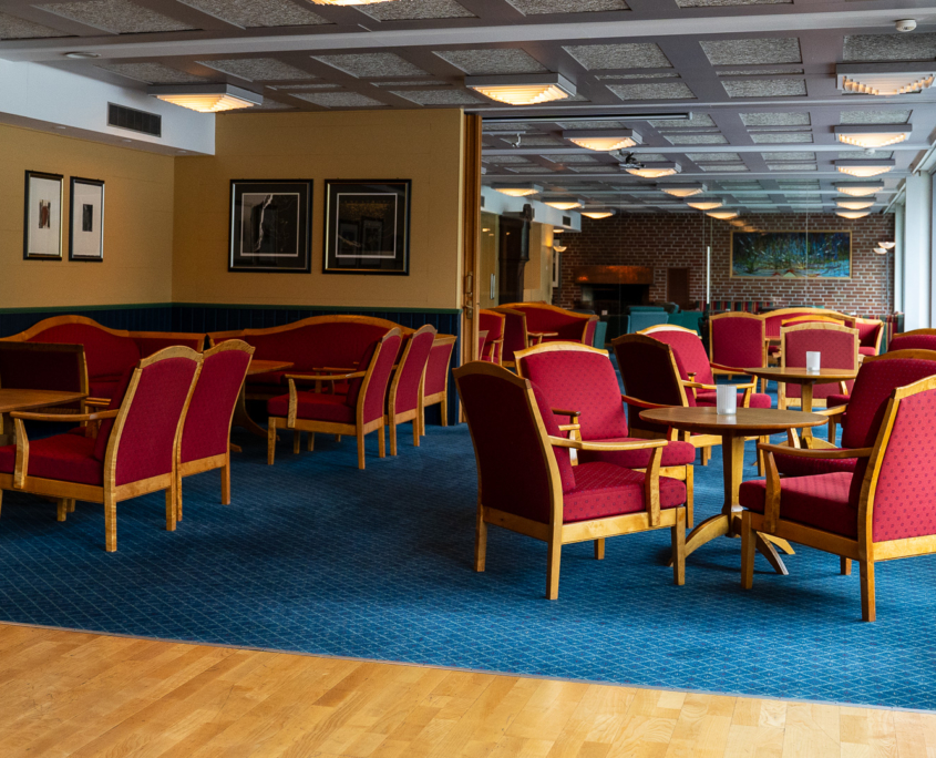 An inviting view of the 7th-floor lounge area at Havila Hotel Geiranger. The space features comfortable seating arrangements and tables, providing a cozy and relaxing atmosphere. The large windows showcase breathtaking views of the fjord, adding a touch of natural beauty to the warm and welcoming ambiance of the hotel lounge.