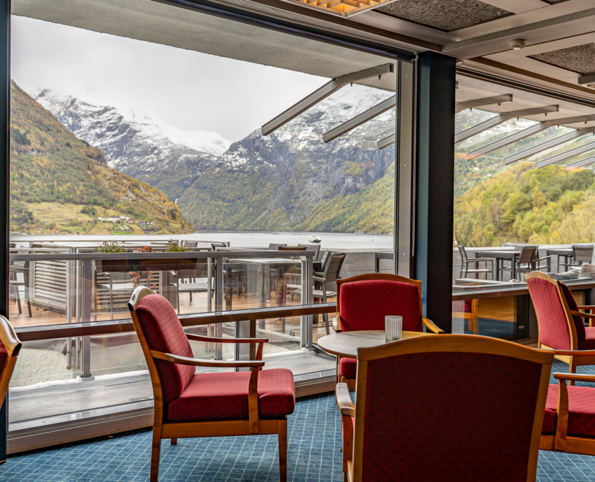 An inviting view of the 7th-floor lounge area at Havila Hotel Geiranger. The space features comfortable seating arrangements and tables, providing a cozy and relaxing atmosphere. The large windows showcase breathtaking views of the fjord, adding a touch of natural beauty to the warm and welcoming ambiance of the hotel lounge.